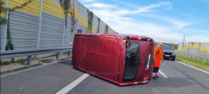 Przewrócony na bok bordowy van na autostradzie, pracownik w pomarańczowym kombinezonie stoi obok, w tle bariery dźwiękochłonne i czarny samochód osobowy.