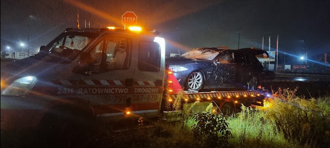 Laweta z uszkodzonym samochodem osobowym w nocnych warunkach, widoczny znak STOP i numer telefonu alarmowego.