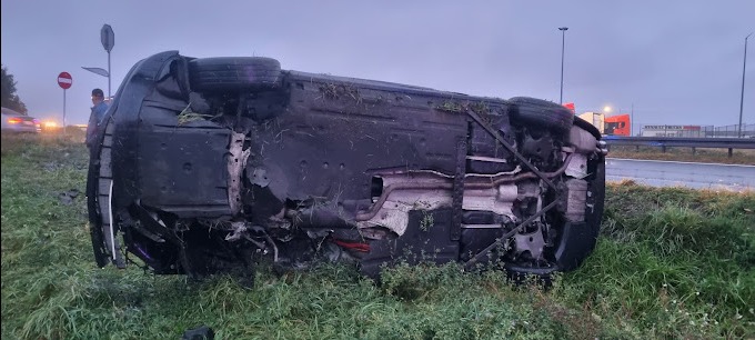 Przewrócony samochód osobowy na poboczu drogi, widoczne uszkodzenia karoserii, w tle znak drogowy zakazu wjazdu i osoba obserwująca zdarzenie.