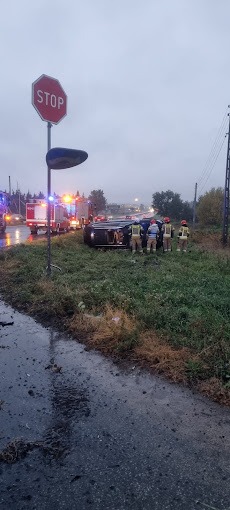 Przewrócony samochód na poboczu drogi, interwencja straży pożarnej, znak STOP w tle, pochmurny dzień.