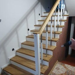 RENO-PAR - Drewniane schody z białymi podstopnicami i balustradą z elementami chromowanymi, widok z dołu, na podłodze płytki ceramiczne z wzorem.