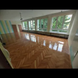 Wnętrze przestronnego pokoju z parkietem ułożonym w jodełkę, po renowacji z widocznym połyskiem, jasne ściany z oknami na zieleń.