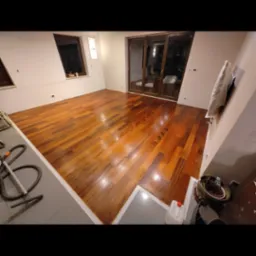 Błyszcząca, świeżo polakierowana podłoga z parkietu w pustym pokoju, widoczny odkurzacz przemysłowy i butelka z preparatem do pielęgnacji podłóg.