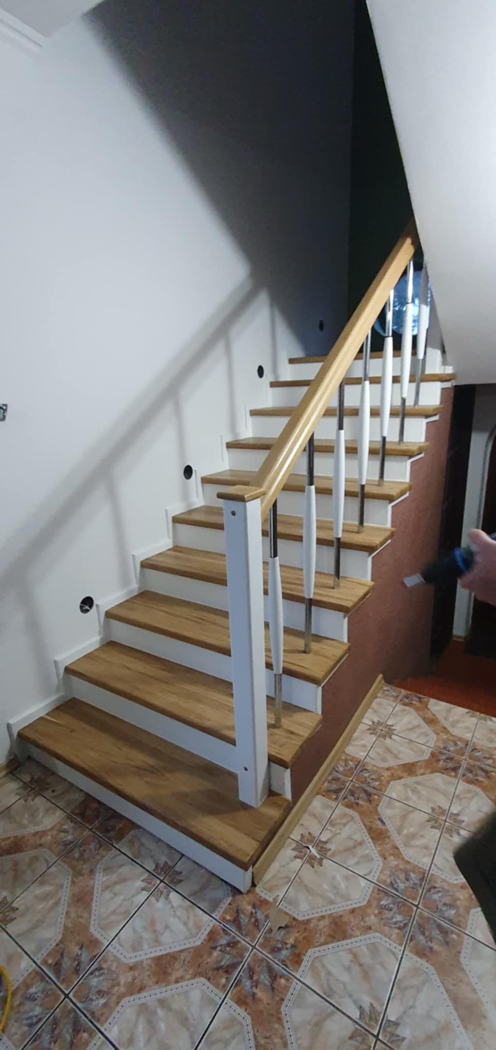 Drewniane schody z białymi podstopnicami i balustradą z elementami chromowanymi, widok z dołu, na podłodze płytki ceramiczne z wzorem.