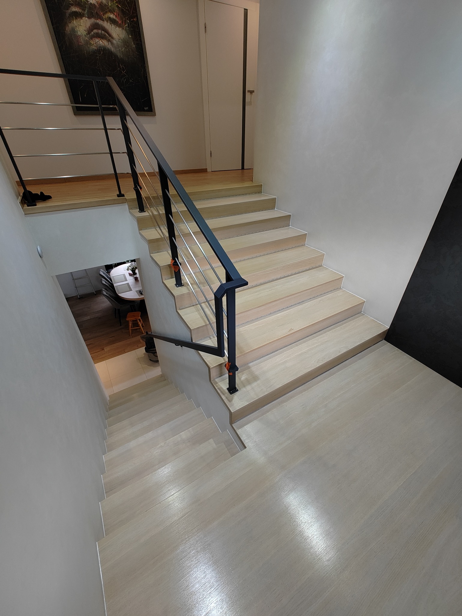 Jasne drewniane schody z czarną metalową balustradą i srebrnymi prętami, widok z góry, w tle obraz na ścianie.