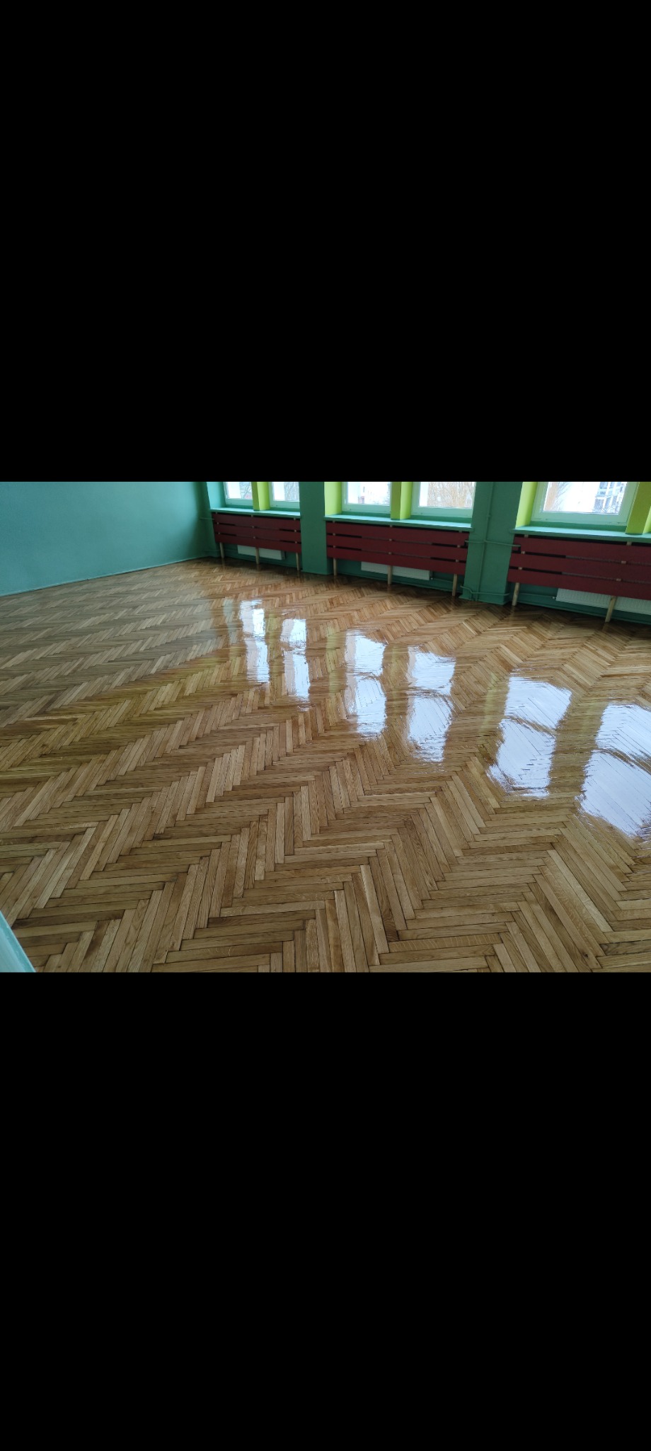 Odnowiona, lakierowana podłoga parkietowa ułożona w jodełkę w przestronnym, jasnym pomieszczeniu z zielonymi ścianami i czerwonymi grzejnikami pod oknami.