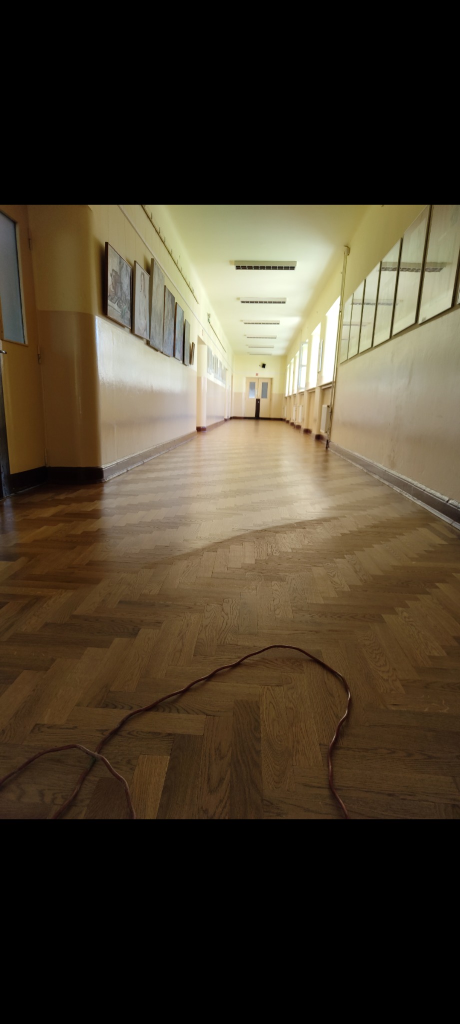 Długi korytarz z parkietem ułożonym w jodełkę, widoczne ślady po cyklinowaniu i kabel na podłodze.