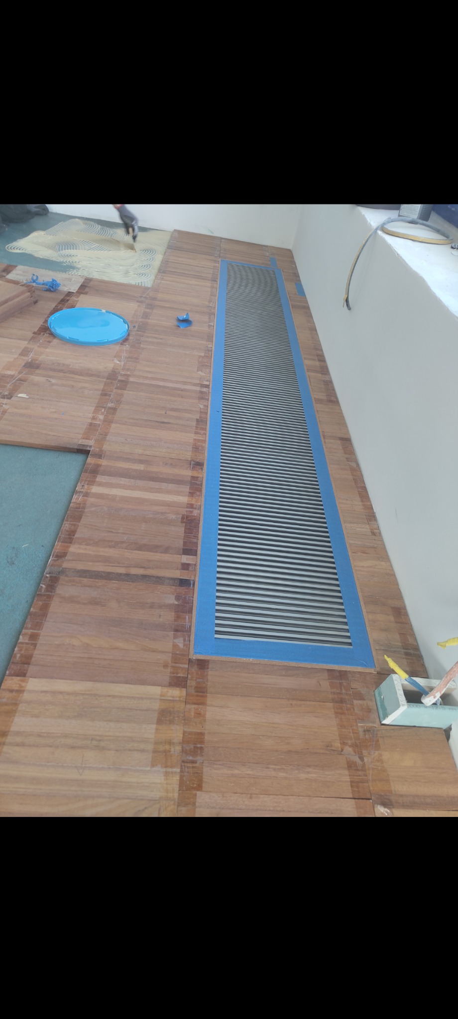 Fragment podłogi z ułożonym parkietem w jodełkę francuską, zabezpieczona taśmą malarską wokół kratki wentylacyjnej, widoczne narzędzia i niebieskie pojemniki.