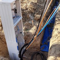 JB Energy Sp. z o.o. - Otwarta skrzynka elektryczna w wykopie ziemnym, widoczne kable w peszlach oraz przewody prowadzone do skrzynki. Wykop zabezpieczony niebieską folią.