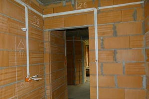 Surowy stan budynku z widocznymi przewodami elektrycznymi prowadzonymi po ścianie z cegły dziurawki, pomarańczowa puszka instalacyjna i wystające przewody, widok na wnętrze przez otwór drzwiowy.