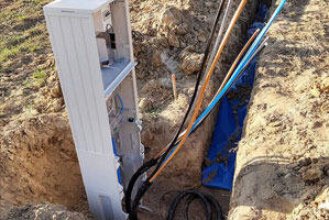 Otwarta skrzynka elektryczna w wykopie ziemnym, widoczne kable w peszlach oraz przewody prowadzone do skrzynki. Wykop zabezpieczony niebieską folią.