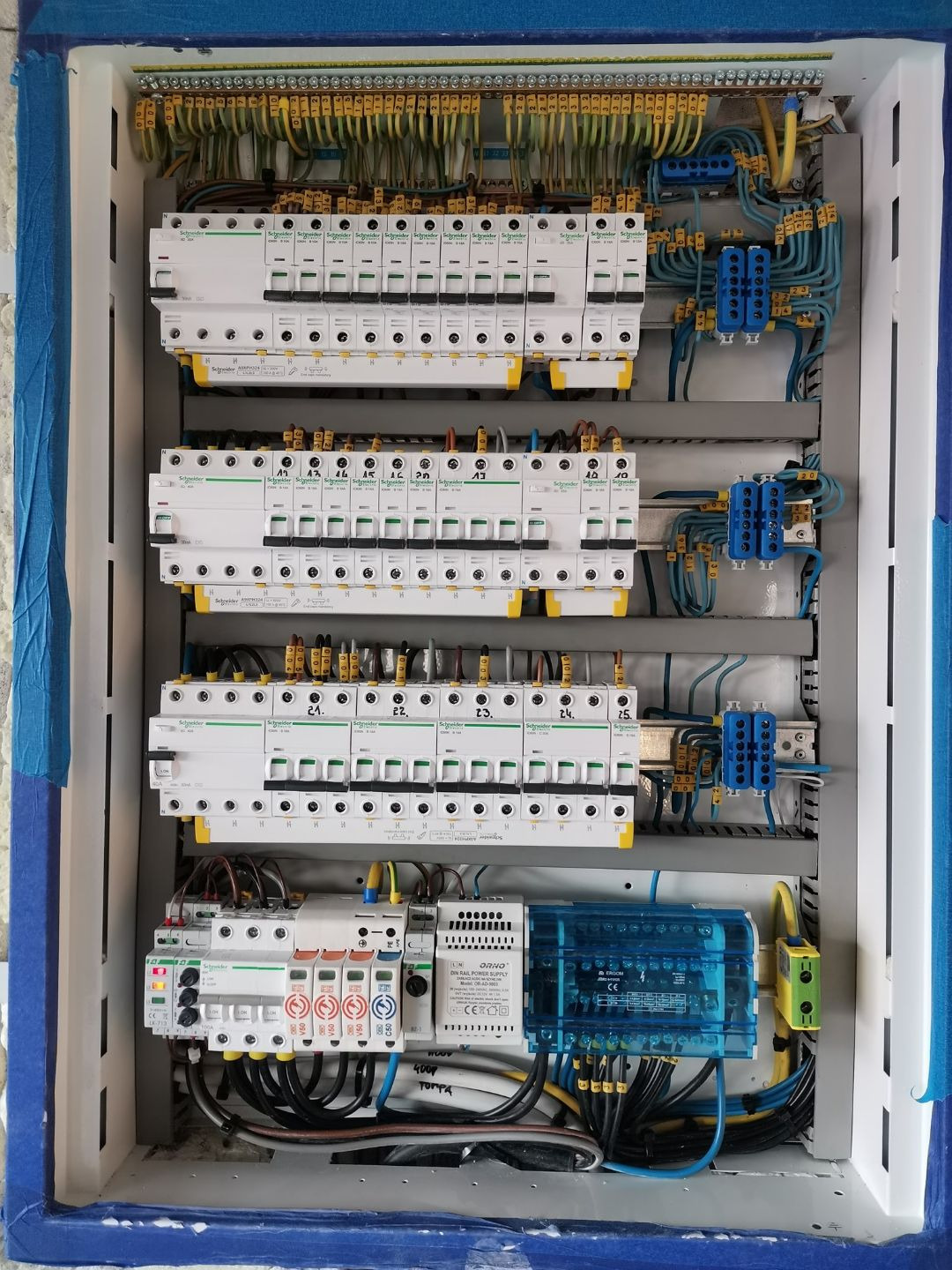 Rozdzielnia elektryczna w trakcie montażu, widoczne zabezpieczenia nadprądowe Schneider, listwy zaciskowe i przewody z oznaczeniami, całość w obudowie z niebieską taśmą malarską na krawędziach.