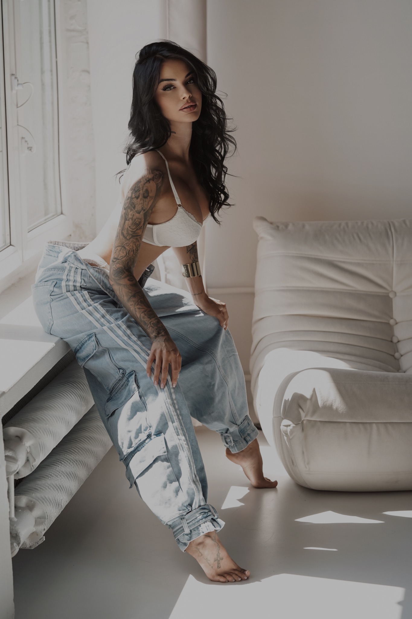 Kobieta z tatuażami na ramieniu, w białym staniku i szerokich jeansach siedzi na parapecie w jasnym wnętrzu, z widocznym fragmentem białego fotela.