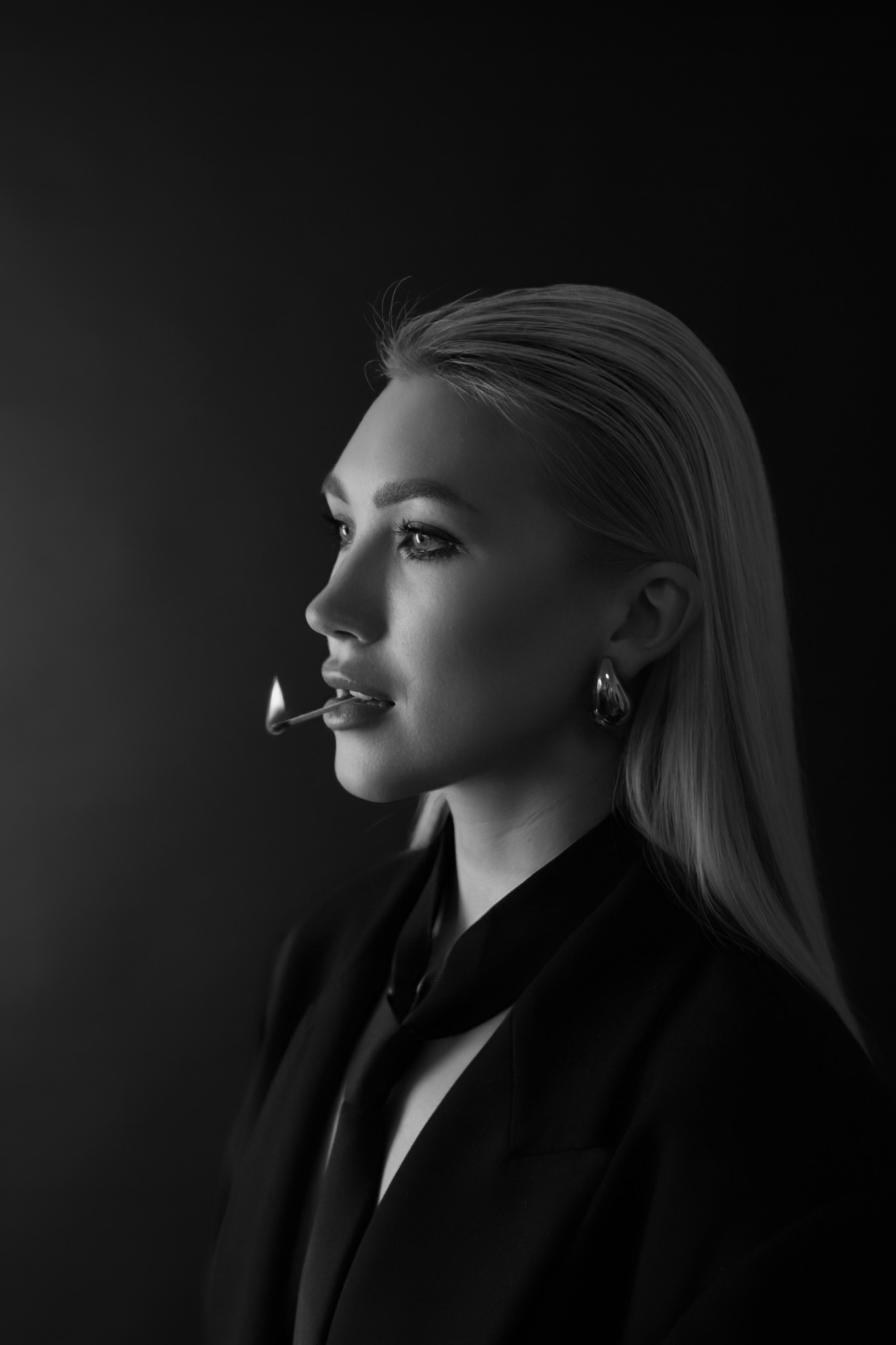 Czarno-białe portretowe ujęcie profilu kobiety z zapaloną zapałką w ustach, w czarnym garniturze i z dużymi kolczykami, na ciemnym tle.