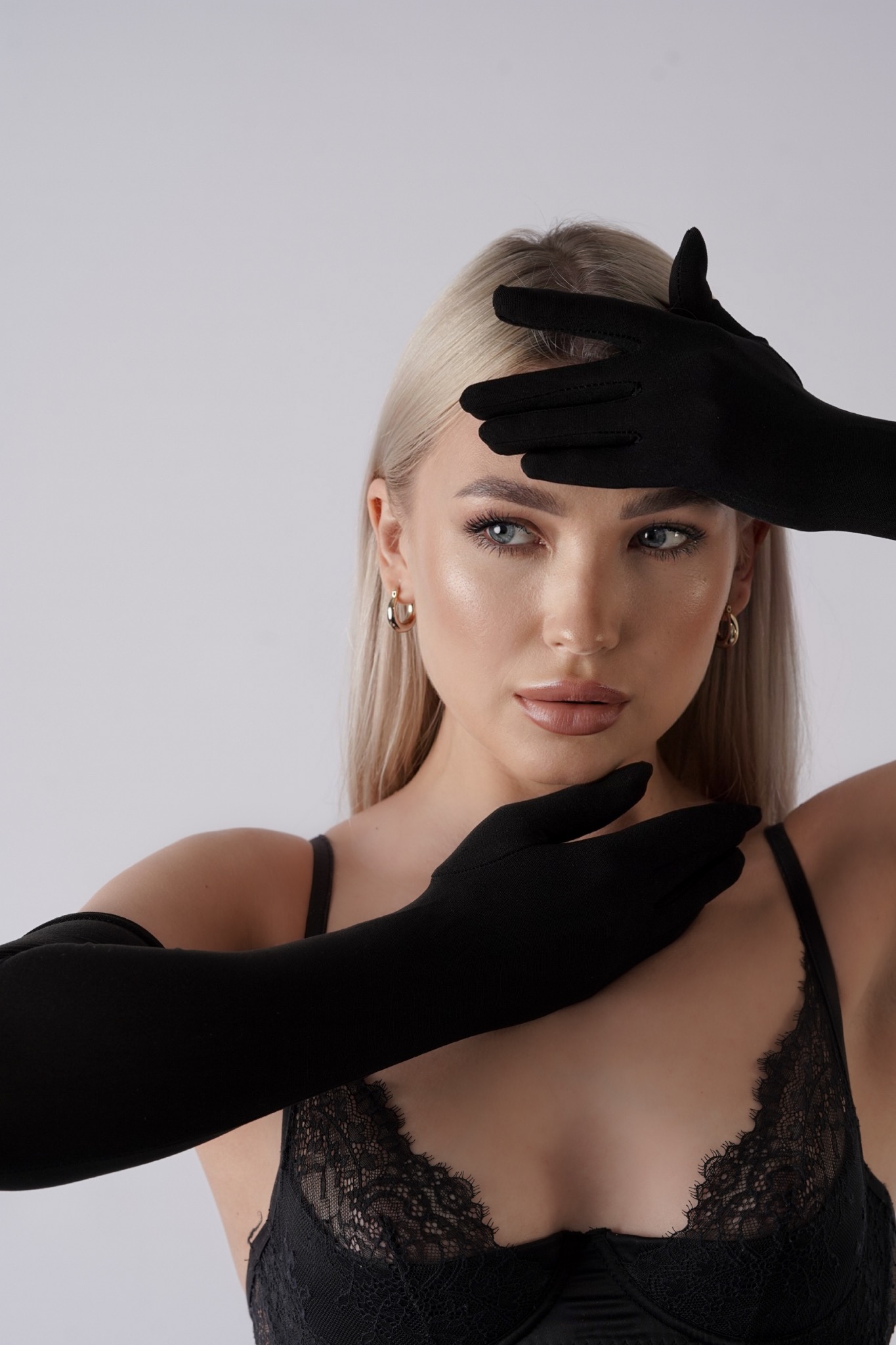 Portret kobiety z blond włosami i niebieskimi oczami, w czarnej koronkowej bieliźnie i długich czarnych rękawiczkach zakrywających twarz.
