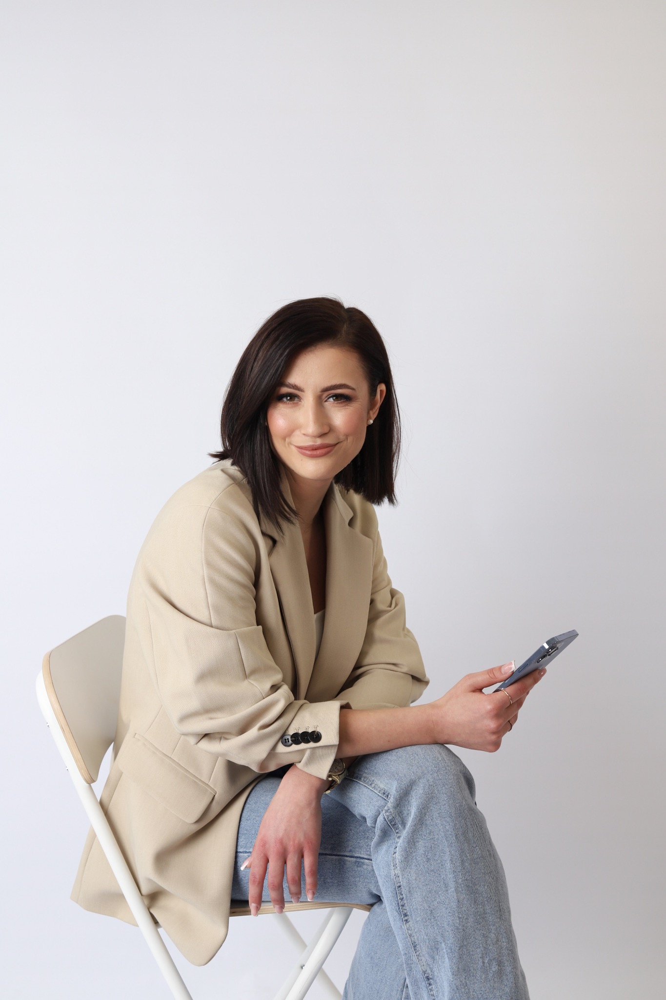 Portret uśmiechniętej kobiety siedzącej na krześle, w beżowej marynarce i jeansach, trzymającej smartfon na białym tle.