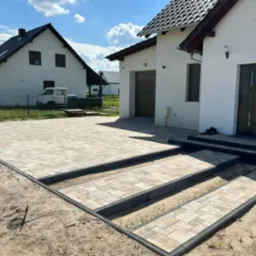 Nowo ułożony, beżowo-szary podjazd z kostki brukowej przed nowoczesnym, białym domem z brązowymi elementami wykończeniowymi; widoczne schody wejściowe z kostki i fragment ogrodu z drugim domem w tle.