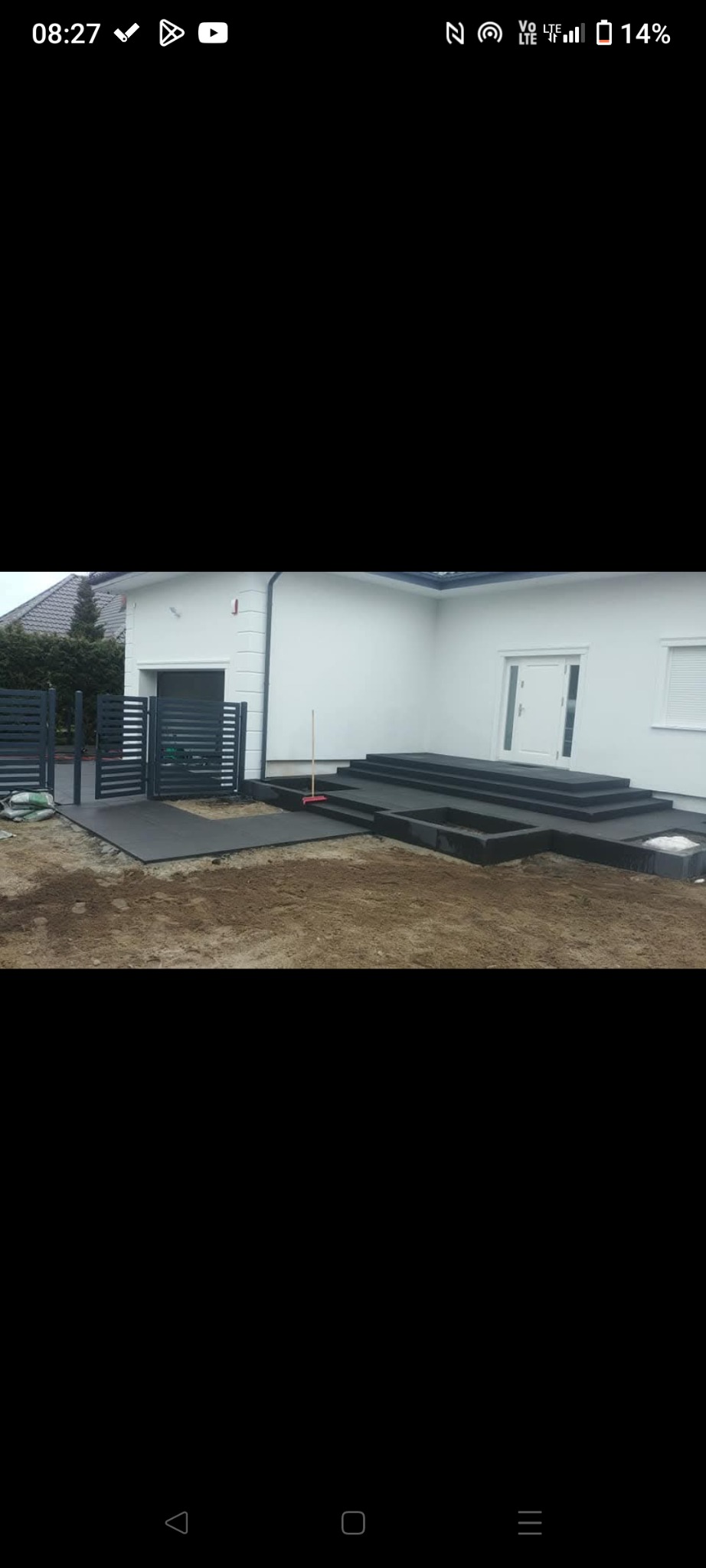 Nowoczesne, antracytowe schody wejściowe i fragment ogrodzenia panelowego przed białym domem, teren częściowo wyrównany, widoczna miotła.