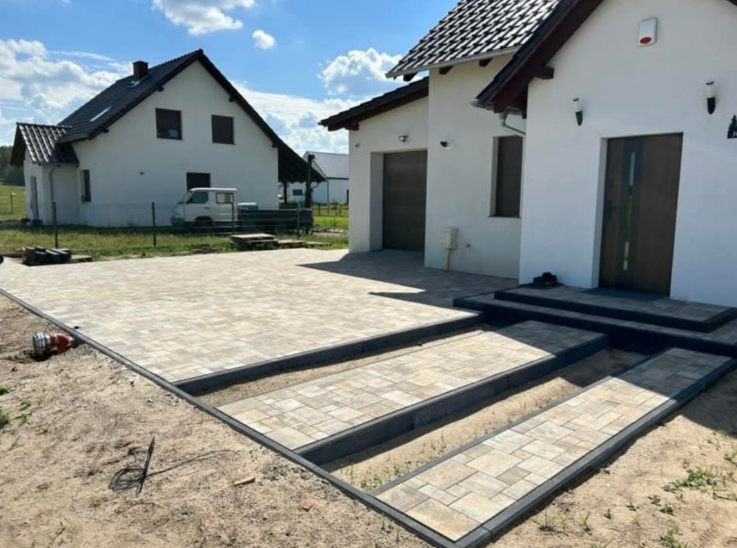 Nowo ułożony, beżowo-szary podjazd z kostki brukowej przed nowoczesnym, białym domem z brązowymi elementami wykończeniowymi; widoczne schody wejściowe z kostki i fragment ogrodu z drugim domem w tle.