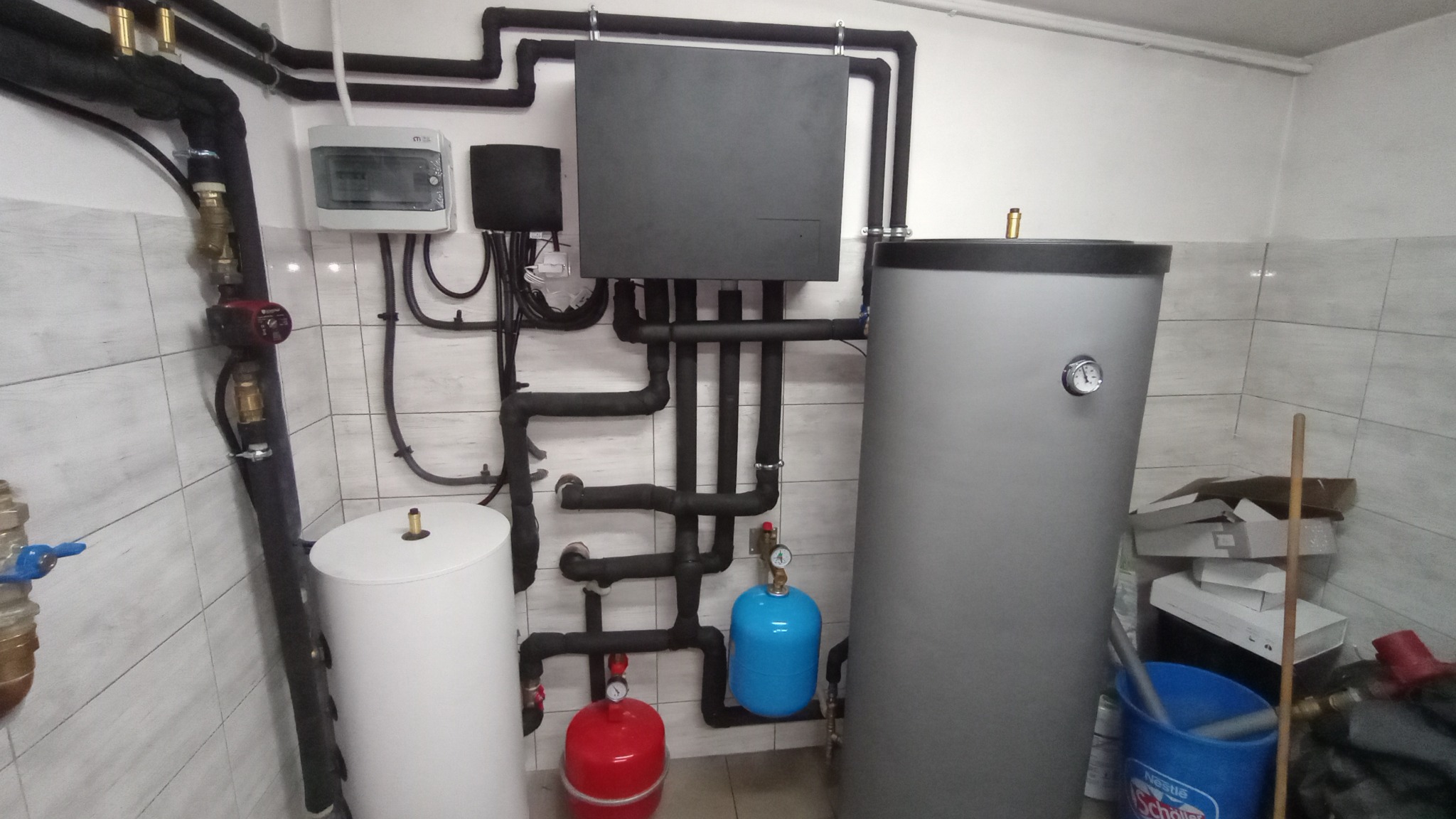 Kotłownia z szarym zbiornikiem buforowym, białym zasobnikiem CWU, niebieskim i czerwonym naczyniem przeponowym, czarnymi rurami instalacji grzewczej i sterownikiem na kafelkowanej ścianie.