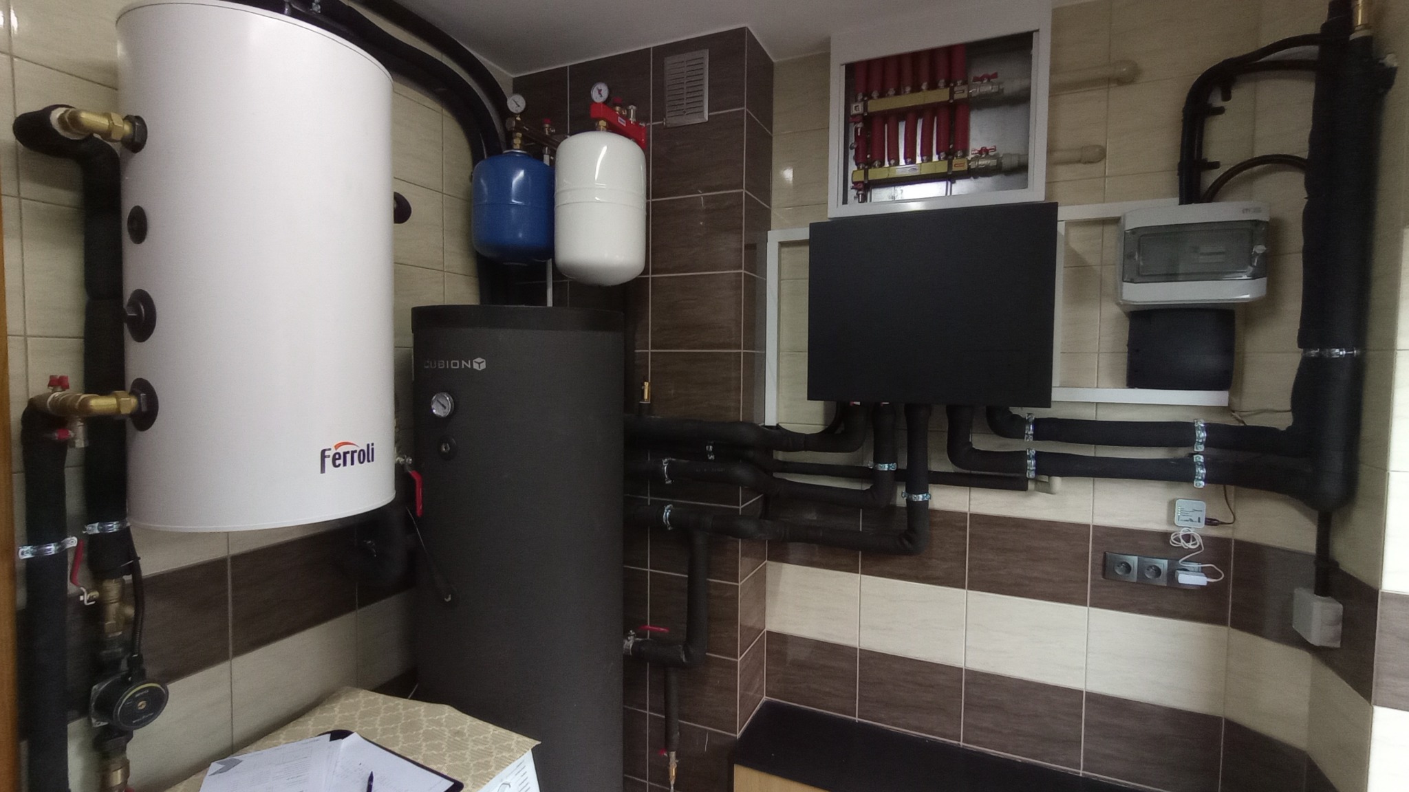 Zestaw urządzeń grzewczych w kotłowni: biały zasobnik ciepłej wody Ferroli, ciemny akumulator ciepła Cubion, naczynia przeponowe, rozdzielacz ogrzewania podłogowego w szafce, orurowanie w izolacji,...