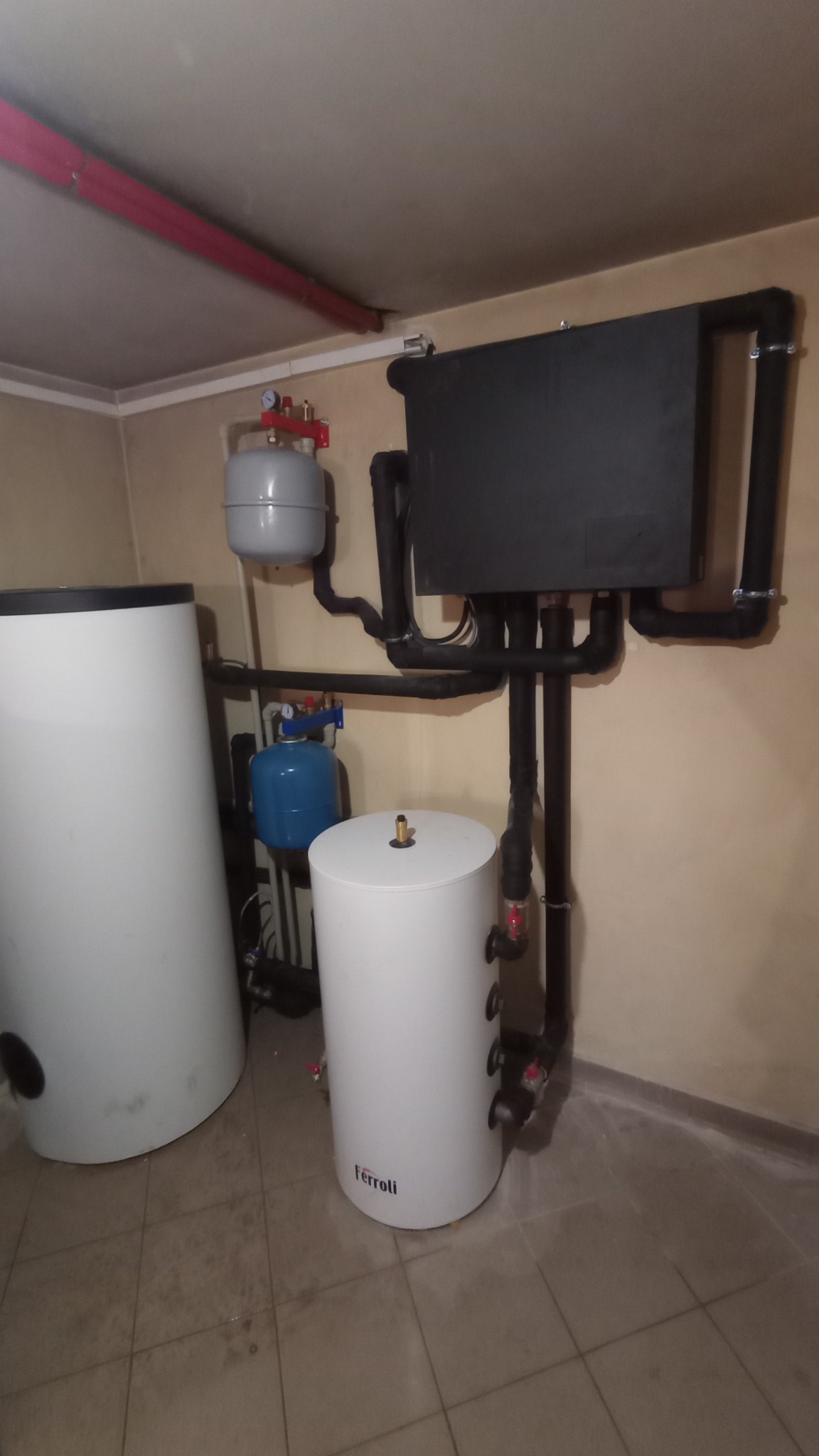 Instalacja centralnego ogrzewania w piwnicy: białe zbiorniki akumulacyjne Ferroli, czarny moduł, rury z izolacją, szare naczynie wzbiorcze i niebieski hydrofor.