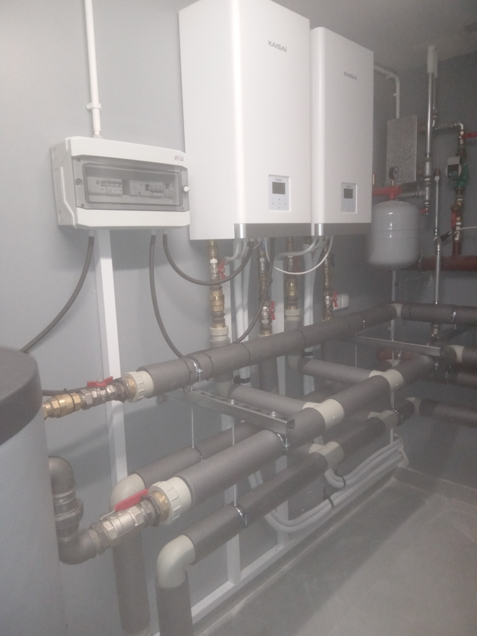 Dwa białe moduły pompy ciepła Kaisai zamontowane na ścianie, z widoczną instalacją rur, zaworów i skrzynką elektryczną, całość w szarej tonacji.
