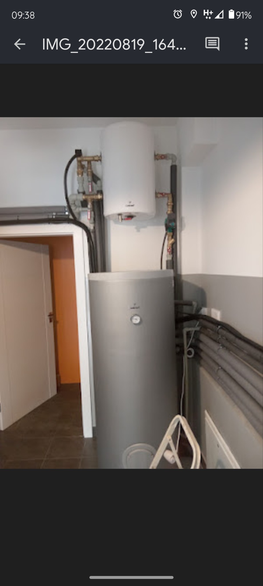 Szary, pionowy zbiornik na wodę z białym, mniejszym zbiornikiem na górze, połączone rurami miedzianymi i czarnymi, oraz licznymi rurami szarymi w tle, widoczne z boku w pomieszczeniu z płytkami.