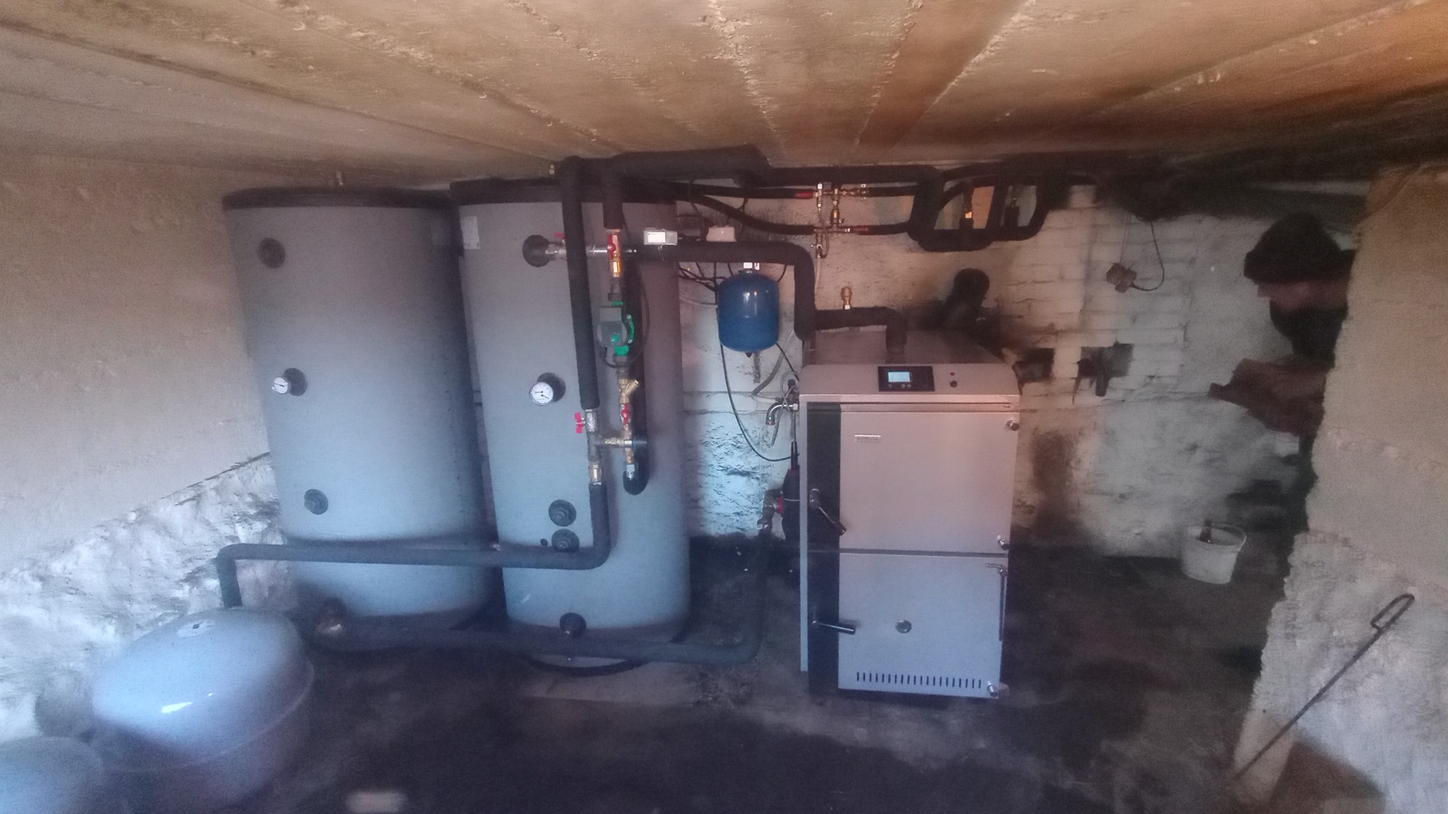 Instalacja grzewcza w piwnicy: dwa szare zbiorniki buforowe, kocioł na paliwo stałe z panelem sterowania i niebieskie naczynie przeponowe, widoczne rury i armatura, osoba w tle.