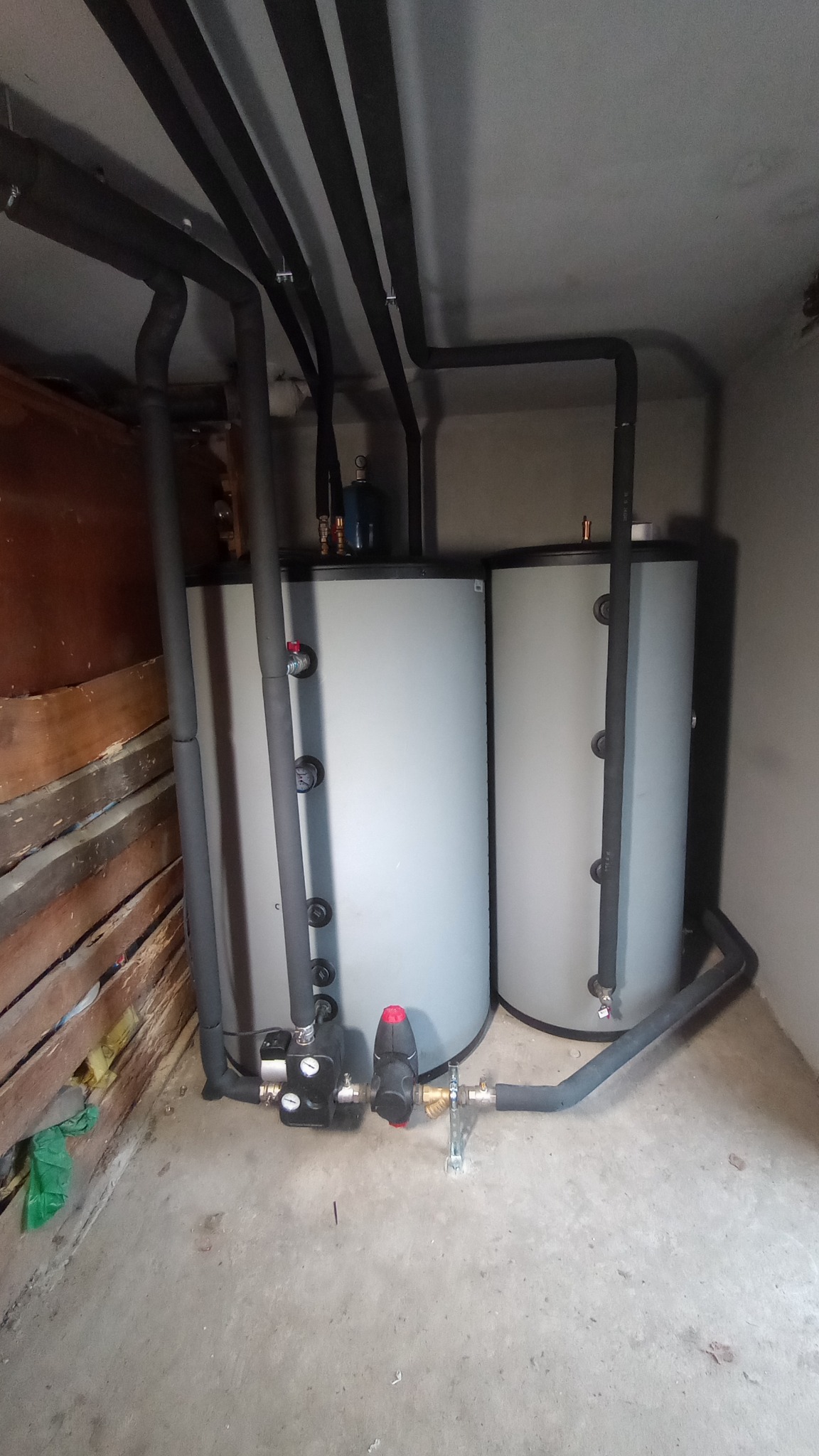 Dwa szare zbiorniki akumulacyjne ciepła z czarnymi rurami w piwnicy w miejscowości Łyse, z widocznymi elementami instalacji i armatury.