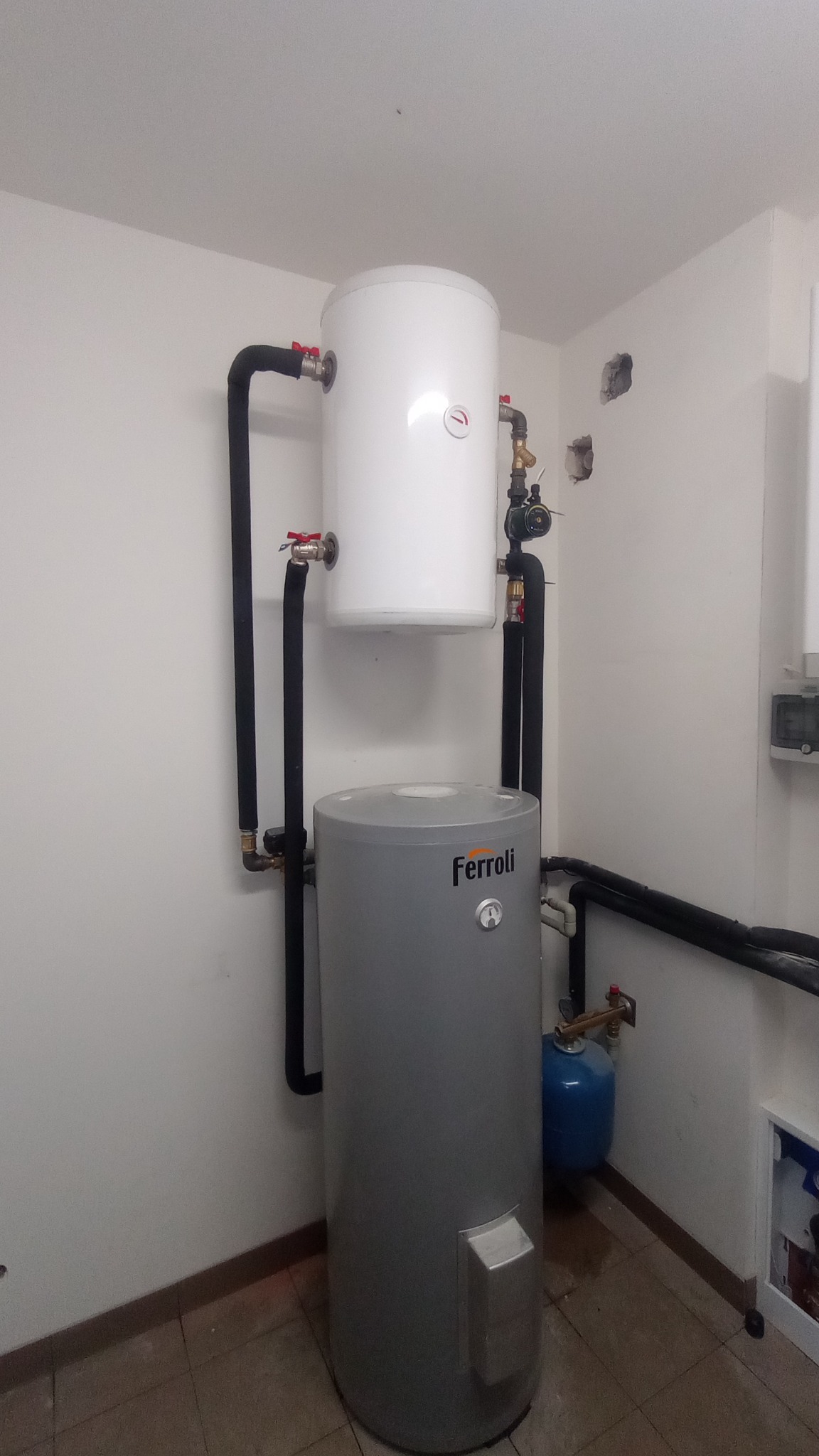 Szary, pionowy zbiornik wody marki Ferroli z manometrem, połączony czarnymi rurami z białym zbiornikiem powyżej, całość zamontowana w rogu pomieszczenia z brązową podłogą z płytek