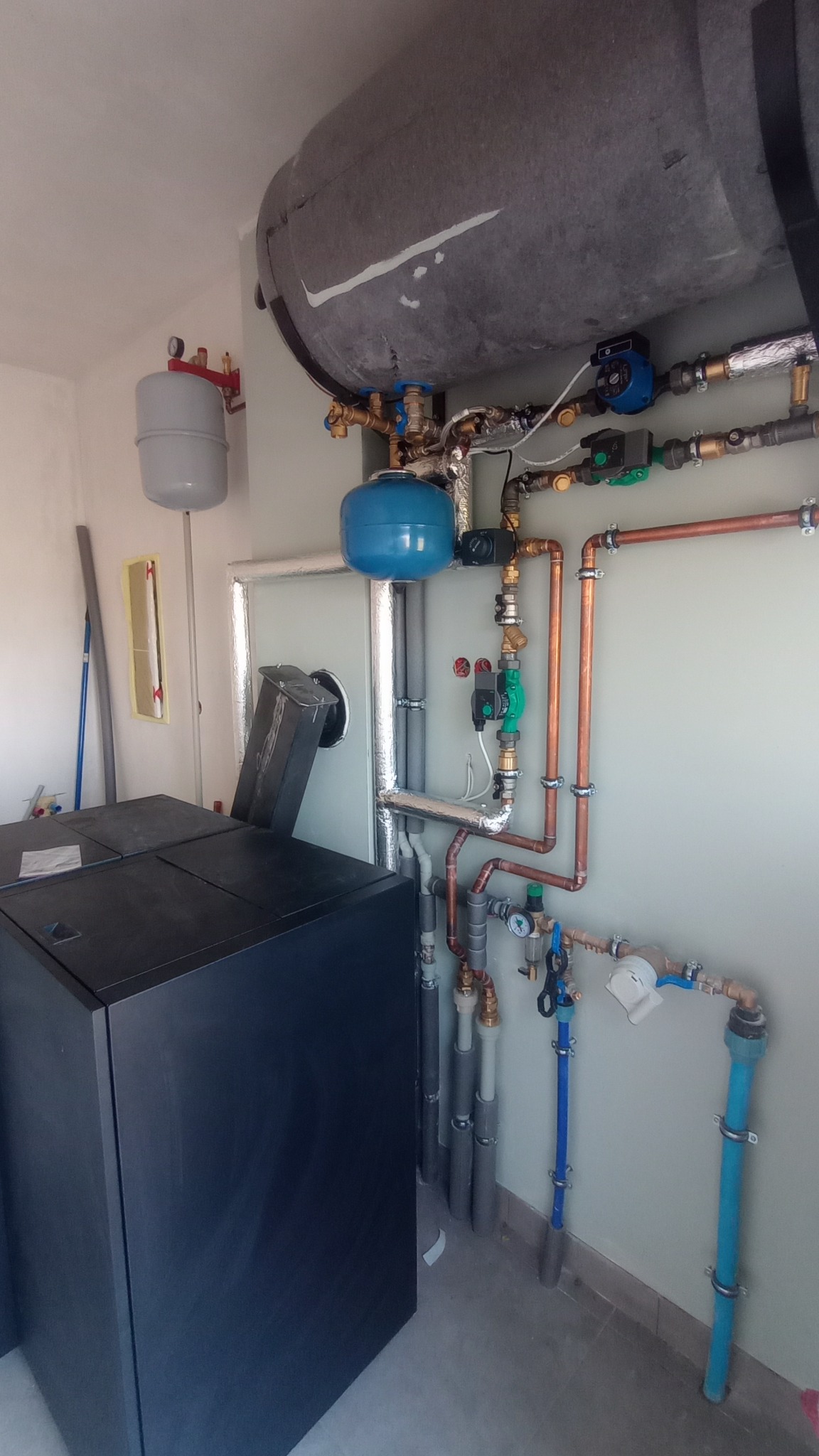 Nowoczesny piec na paliwo stałe z podłączeniami hydraulicznymi: rury miedziane, zawory, pompy obiegowe, zbiorniki wyrównawcze. Widoczne elementy instalacji grzewczej w kotłowni.