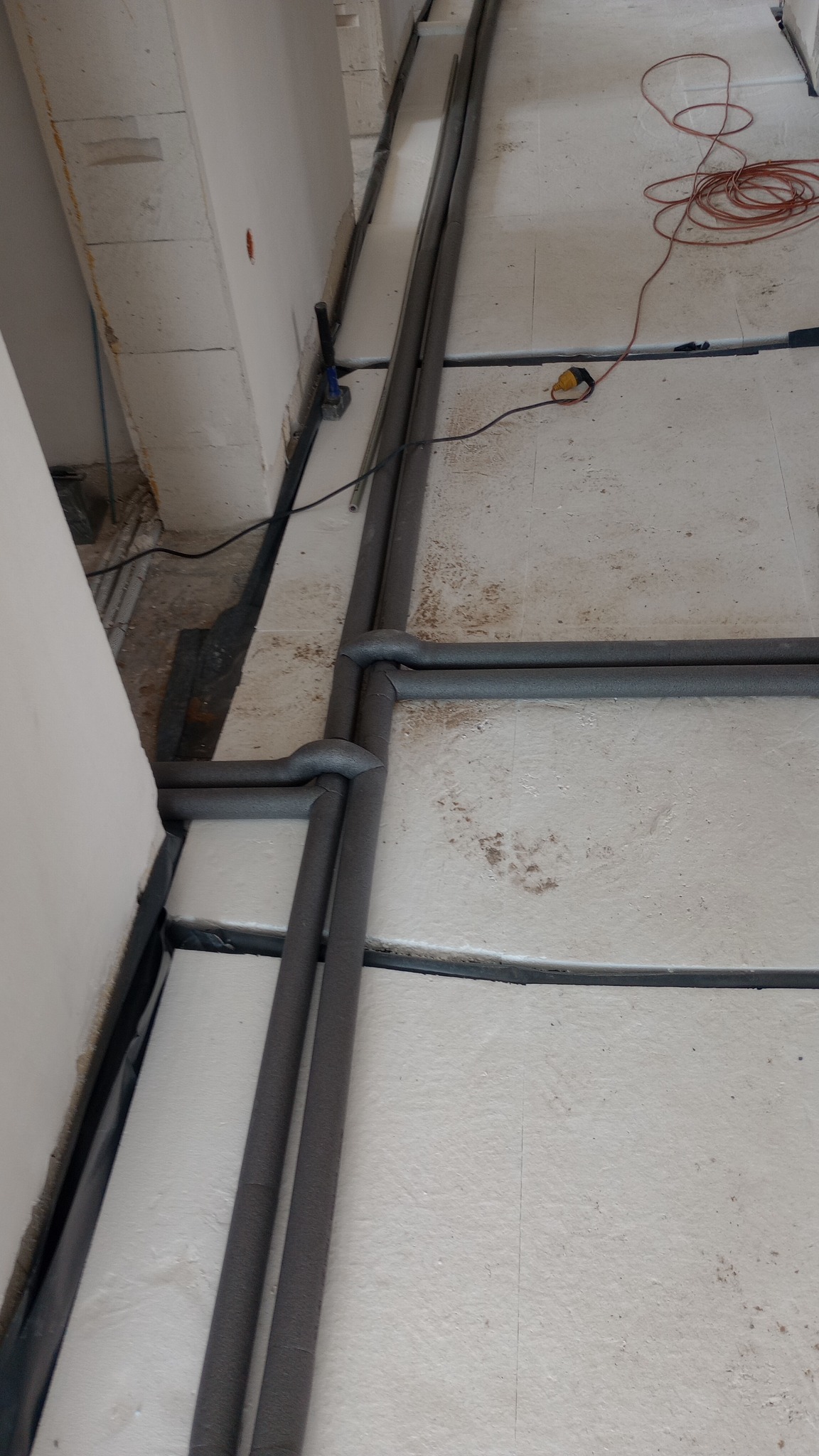 Izolowane rury grzewcze ułożone w bruzdach w podłodze podczas prac instalacyjnych, widoczny kabel zasilający i narzędzia.