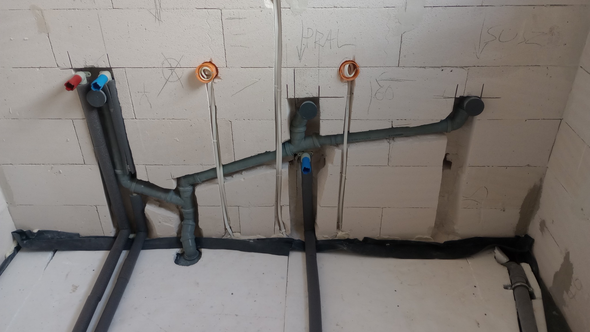 Szare rury kanalizacyjne i wodne w trakcie montażu w surowym wnętrzu z białych bloczków, widoczne otwory na gniazdka elektryczne i instalacje, czarna izolacja przy podłodze.