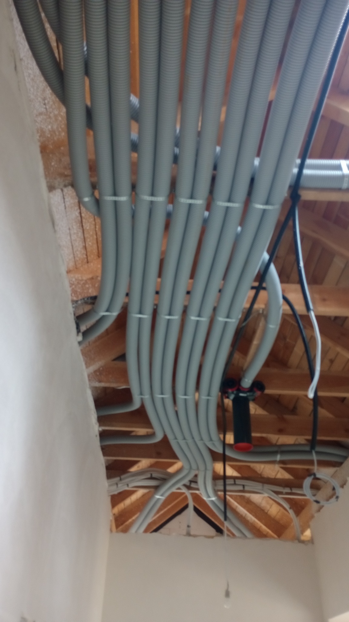 Szereg szarych, karbowanych rur instalacyjnych, przymocowanych białymi opaskami do drewnianej konstrukcji stropu poddasza, widoczne elementy instalacji elektrycznej i hydraulicznej.