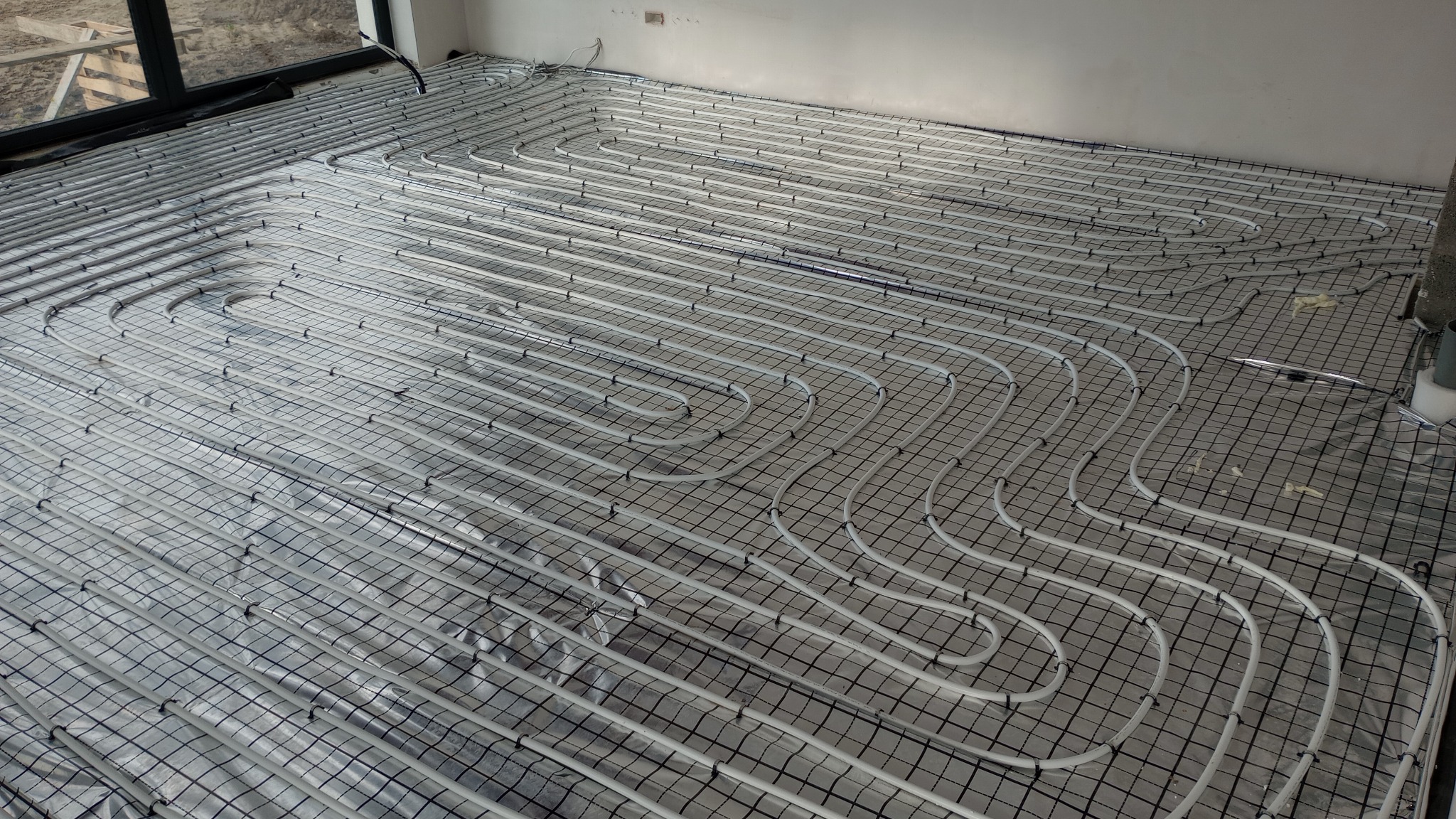 Rozłożona instalacja ogrzewania podłogowego z białych rurek na srebrnej folii izolacyjnej z czarną siatką, widoczne okno i fragment ściany.