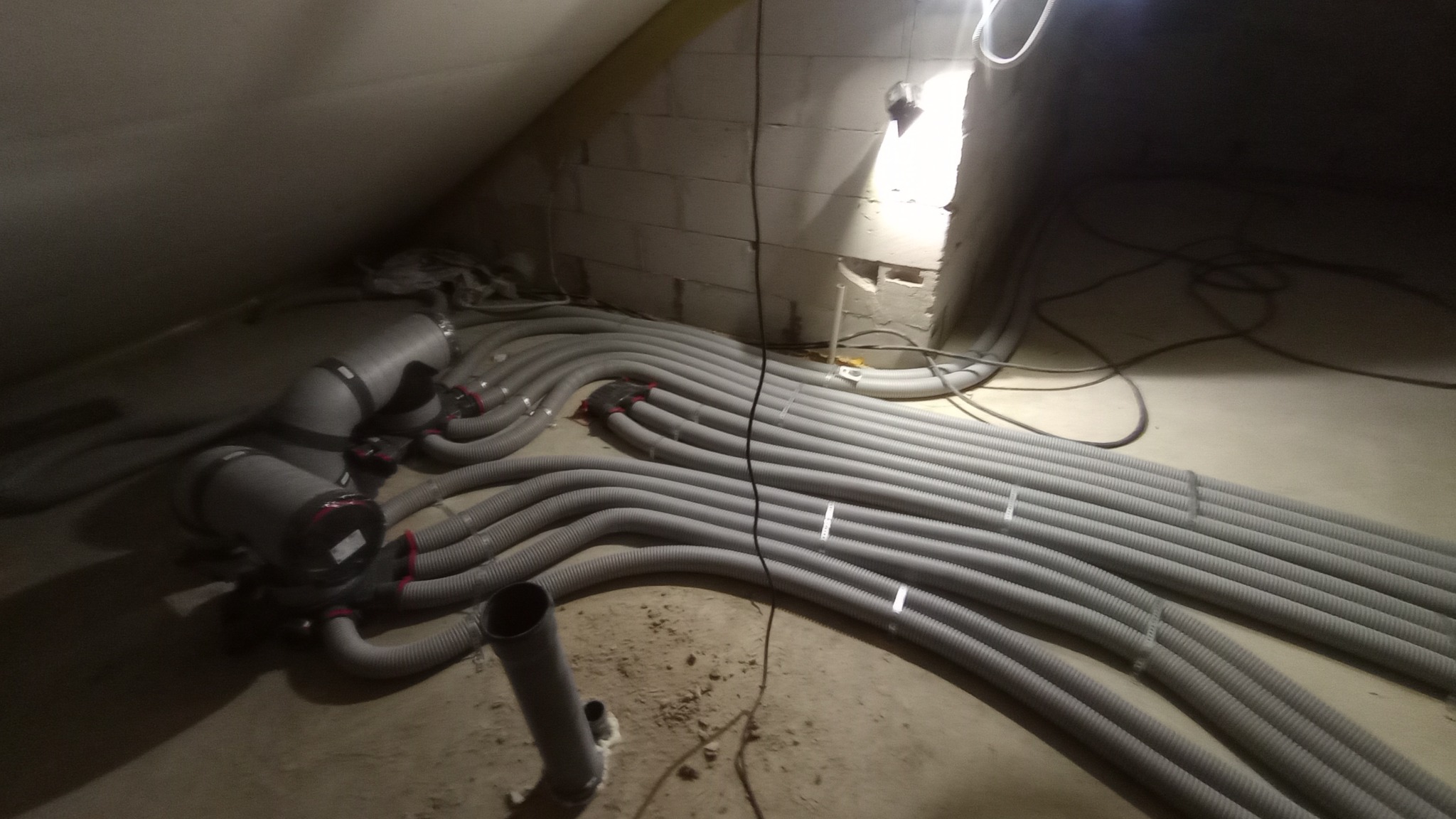 Szare, elastyczne rury wentylacyjne ułożone równolegle na podłodze poddasza, połączone z rozdzielaczem, widoczne przewody elektryczne, surowe ściany z betonu komórkowego.