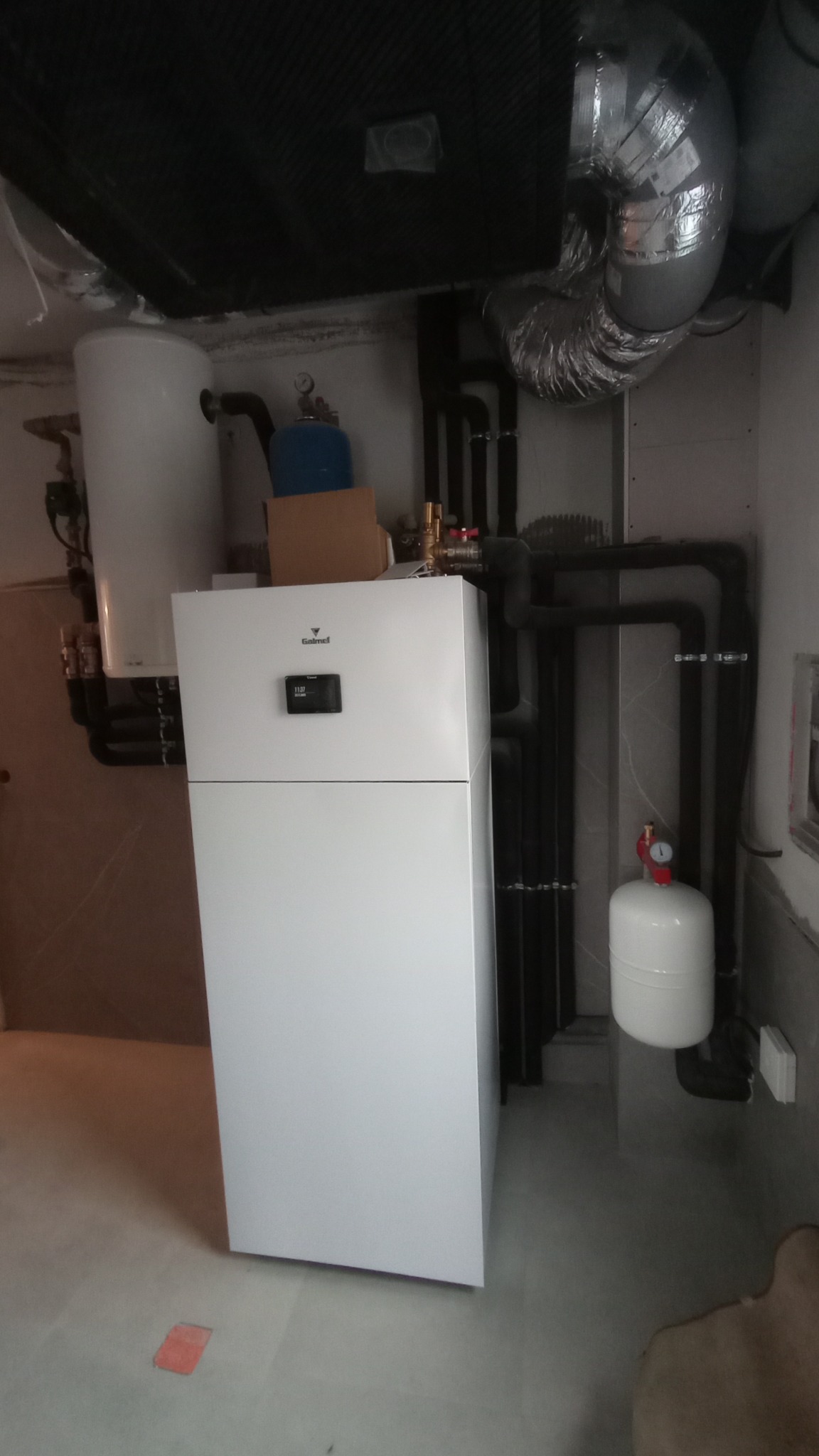 Biała pompa ciepła Galmet w kotłowni, widoczne rury instalacyjne, zbiornik wyrównawczy i elementy wentylacji pod sufitem.