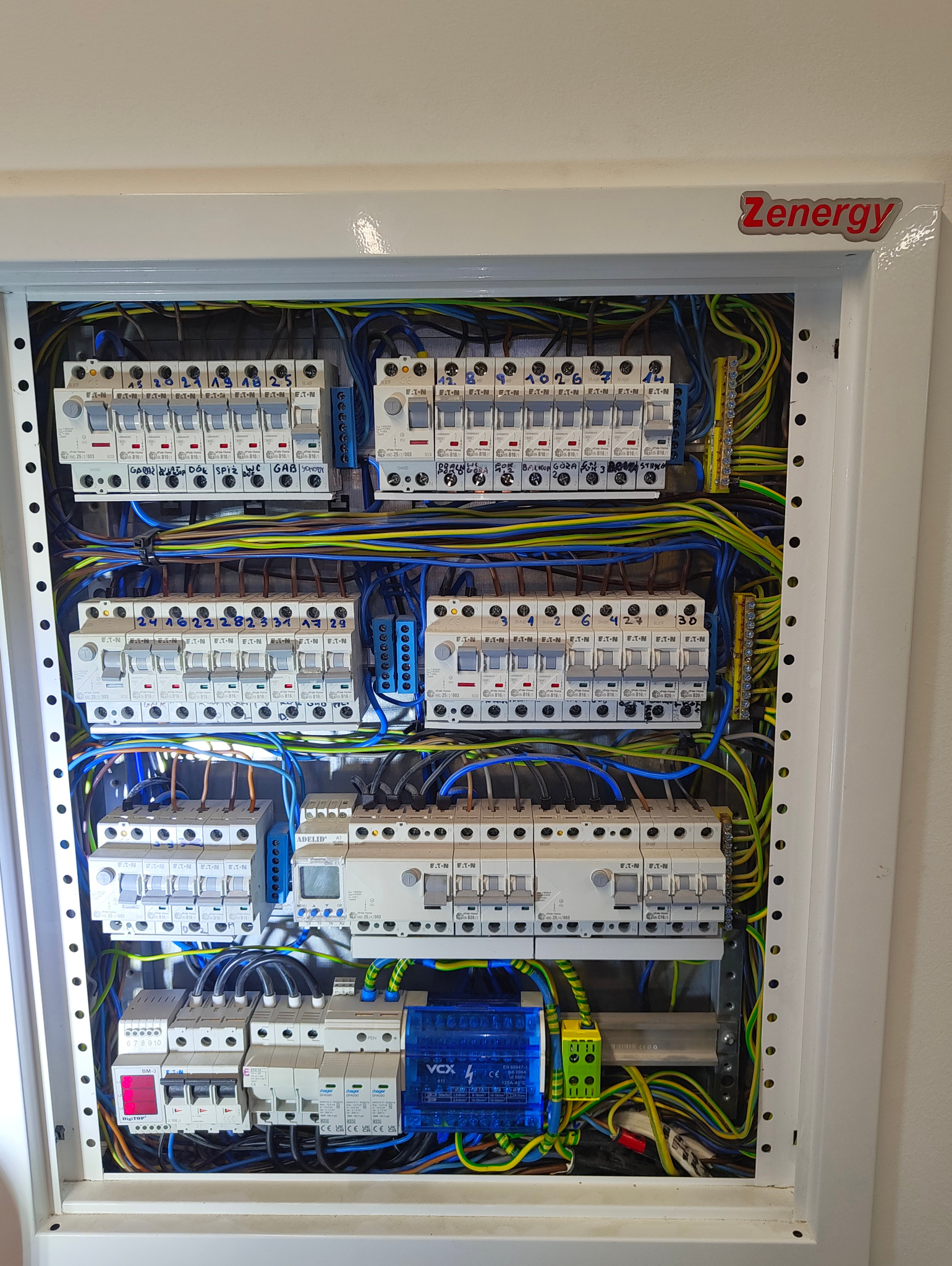 Rozdzielnica elektryczna w białej obudowie, widoczne liczne bezpieczniki Eaton i Hager, przewody w kolorach niebieskim, żółto-zielonym i czarnym, opisane obwody, w dolnej części moduł VCX 4.