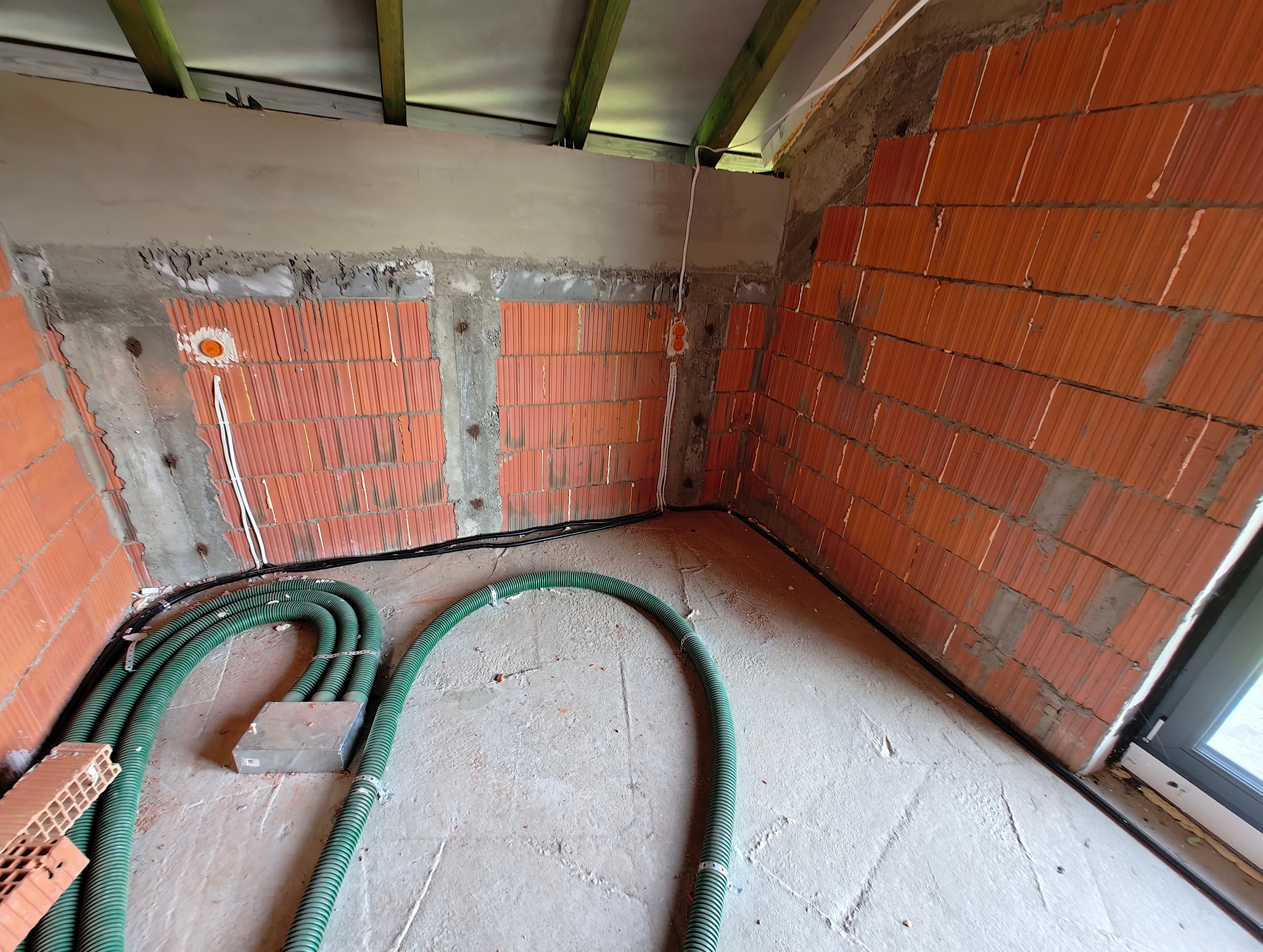 Wnętrze w trakcie prac: ściany z czerwonej cegły z widocznymi puszkami instalacyjnymi i przewodami elektrycznymi w zielonych karbowanych rurach ułożonych na surowej posadzce.