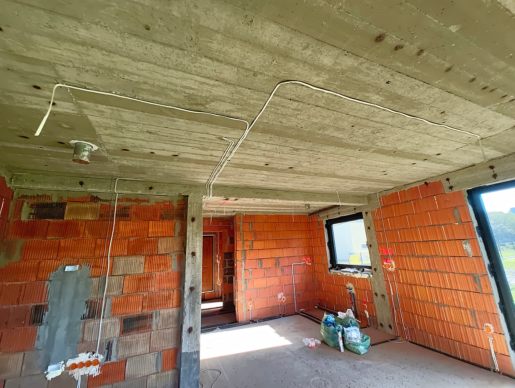 Surowy stan domu jednorodzinnego w trakcie rozprowadzania przewodów elektrycznych podtynkowo po suficie i ścianach z czerwonej cegły, widoczne puszki instalacyjne i worki z materiałami budowlanymi.