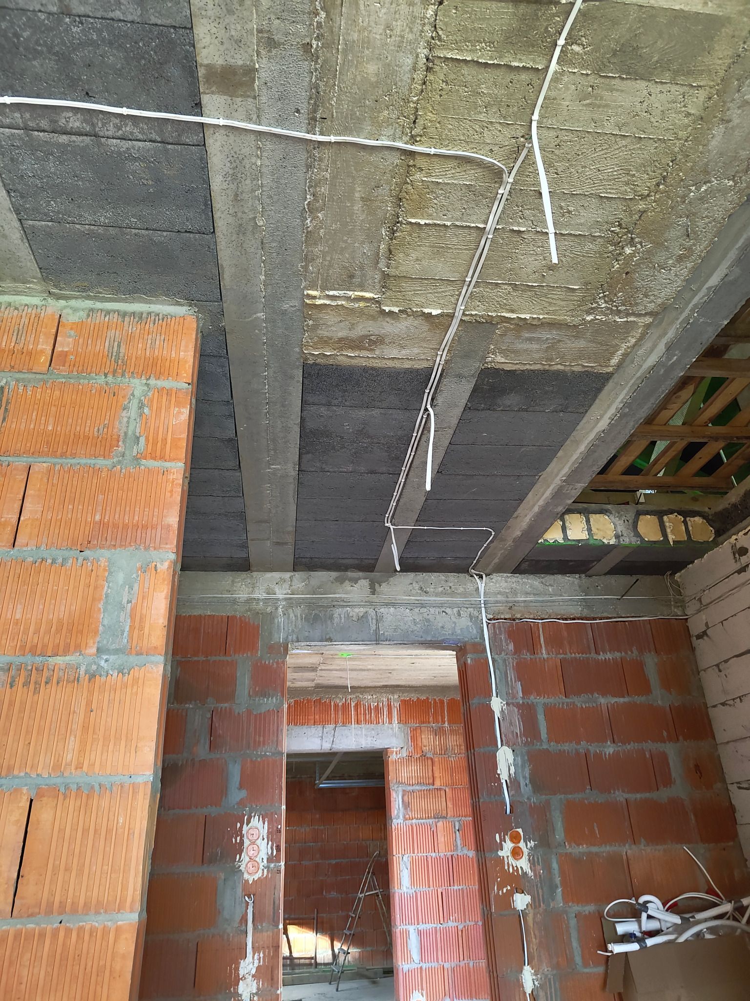 Surowy stan budynku w trakcie prac: widoczne przewody elektryczne przymocowane do sufitu i ścian z cegieł, z otworami pod gniazdka elektryczne i puszkami instalacyjnymi.