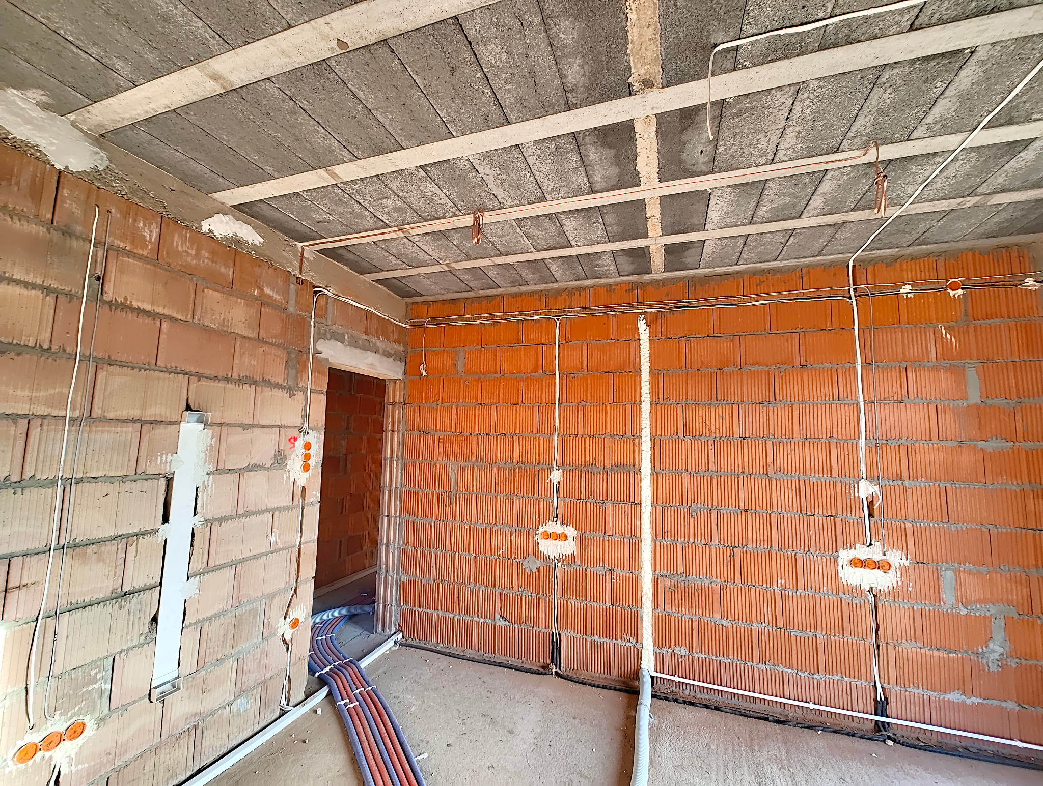 Surowy stan budynku w trakcie prac. Widoczne kable elektryczne prowadzone po ścianach z czerwonej cegły dziurawki, puszki instalacyjne częściowo zamurowane, rury instalacyjne w podłodze, sufit...