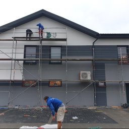 Usługi Malarsko-Budowlane - Dwóch pracowników na rusztowaniu maluje elewację budynku na biało i szaro, trzeci pracownik na dole przygotowuje farbę, widoczne zabezpieczone okna i klimatyzator na ścianie.