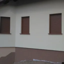 Fragment ocieplonego budynku z brązowymi roletami zewnętrznymi w oknach i rynną spustową. Widoczny cokół budynku w dwóch odcieniach brązu.