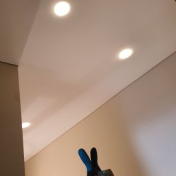 Usługi Malarsko-Budowlane - Gładka, jasna powierzchnia sufitu z trzema wpuszczonymi punktami świetlnymi, widok z dołu, z fragmentem ściany i narzędziami malarskimi w rogu kadru.