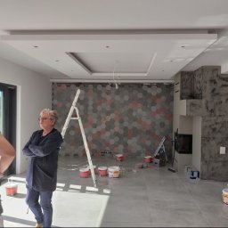 Usługi Malarsko-Budowlane - Wnętrze w trakcie wykańczania: ściana z heksagonalnych płytek w odcieniach szarości i czerwieni, drabina aluminiowa, kobieta z założonymi rękami obserwująca postęp prac, puszki z farbą na podłodze,...