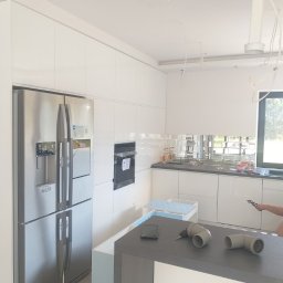 Usługi Malarsko-Budowlane - Montaż nowoczesnej, białej kuchni w Budzyniu: widoczna lodówka side-by-side, zabudowany piekarnik, szklane płytki nad blatem oraz elementy instalacji wodno-kanalizacyjnej na wyspie kuchennej.