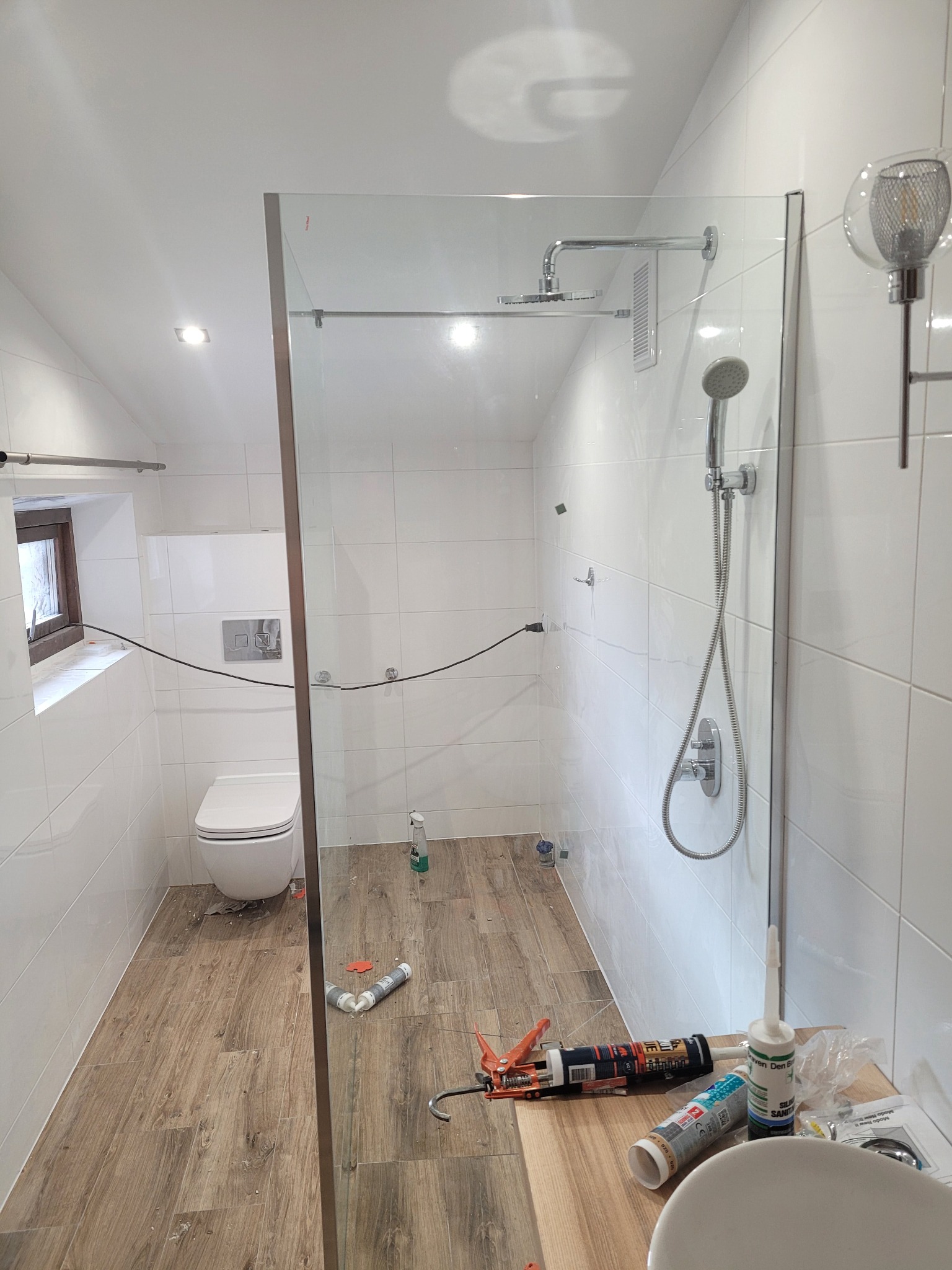 Nowoczesna łazienka w trakcie wykańczania: kabina prysznicowa ze szklaną ścianką, białe płytki na ścianach, drewnopodobne panele na podłodze, widoczne narzędzia i materiały budowlane, lampa w stylu...