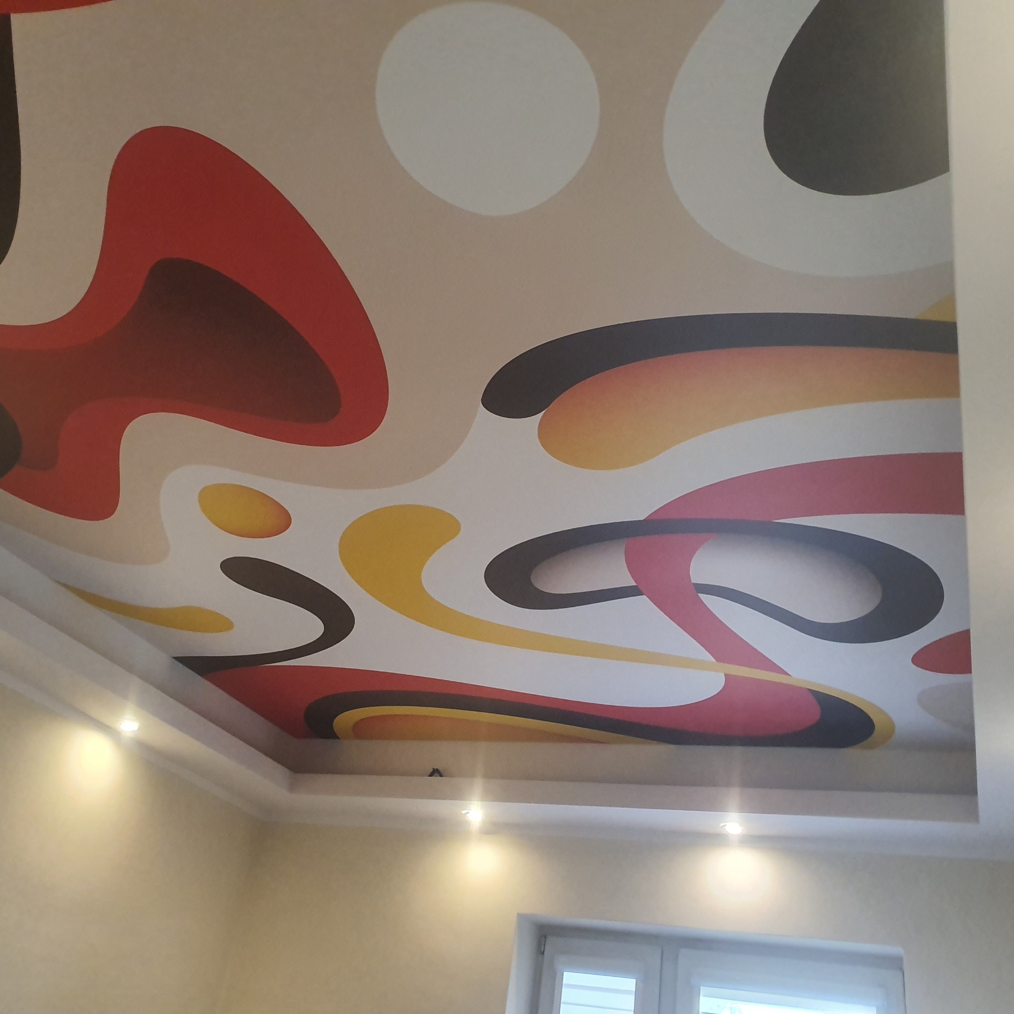 Sufit napinany z abstrakcyjnym wzorem w kolorach czerwonym, żółtym, czarnym i białym, oświetlony punktowymi lampami.